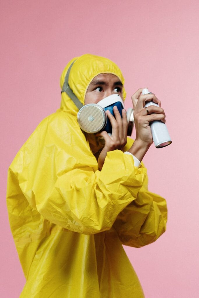 A person in a yellow hazmat suit holding a spray can against a pink background.